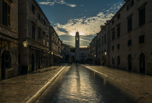 Ein Kirchenturm in Dubrovnik blockiert die Sonne am Morgen, die Straße ist leer