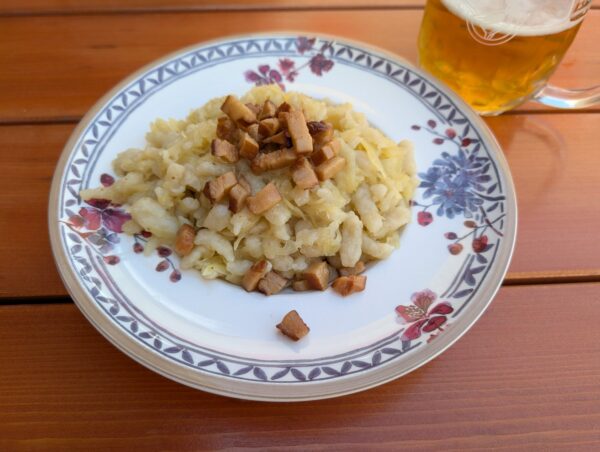 Ein Teller mit Nockerln, Kraut und gebratenem Bauchspeck und ein Glas Bier auf einem Holztisch